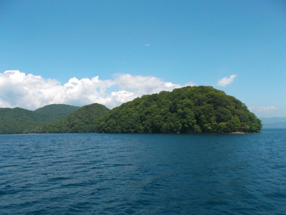 We see a clear blue sky above, with some low lying white clouds in the distance.  In the center we see forested islands in the lake.  The bottom half of the picture is all blue, blue lake.