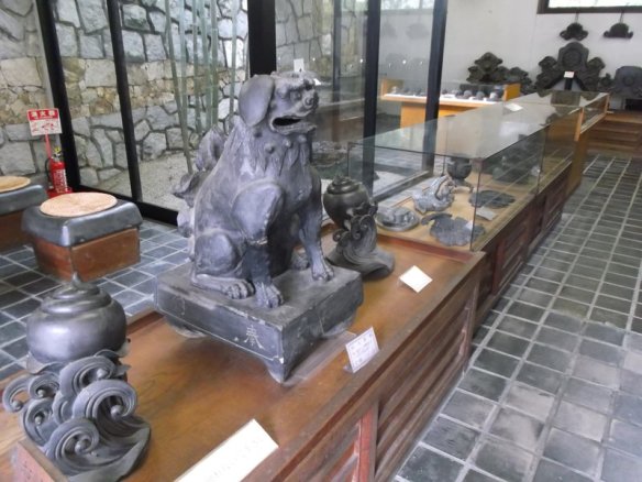 The photo shows a statue of a puppy sitting upright on its hind legs carved in stone, in a room with other sculptures with glass cases and stonework on the walls and floors