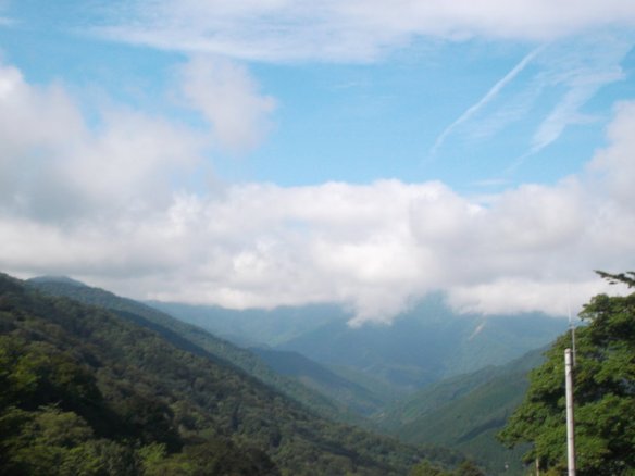 Above is a blue sky with little white puffs of clouds.  Below is a valley, flanked by green mountains on both sides, heading straight into another green mountain in the distance.  Above the distant green mountain is a blanket of white clouds