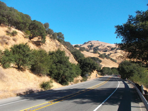 Under a big blue sky, we see hills covered with yellow dead grass with splotches of green trees on them, and a road winds around the hills in the bottom right